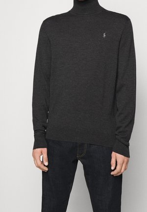Homme portant un pull à col roulé gris foncé avec un petit logo brodé sur la poitrine et un jean bleu foncé, debout devant un fond uni.