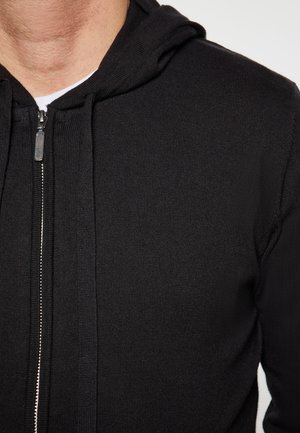 Sort zip-up hættejakke med ribbet struktur, der har en metallynlås og en hætte. Stoffet ser blødt og strikket ud.