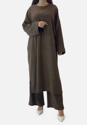Mujer que lleva un vestido marrón de manga larga con textura acanalada sobre pantalones de pierna ancha a juego, con tacones blancos de punta afilada, de pie frente a un fondo liso.