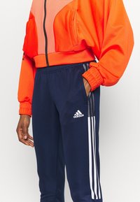 Orange cropped jacka med en texturerad panel, tillsammans med marinblå byxor med vita ränder och en Adidas-logotyp på sidan.