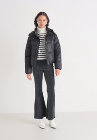 Schwarze Steppjacke mit hohem Kragen, horizontale Streifen auf einem weißen Rollkragenpullover, glitzernde schwarze Schlaghosen und weiße Knöchelstiefel.