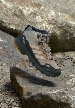 Beiger und schwarzer Wanderstiefel mit Gore-Tex, Terrex-Branding und grober Sohle, der über felsigem Gelände schwebt.
