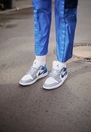 Osoba nosząca białe i niebieskie sneakersy Nike Air Jordan z szarymi sznurowadłami i niebieskie spodnie stoi na asfalcie obok pokrywy studzienki.
