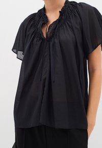 Blouse noire transparente avec col à volants, manches courtes évasées et fermeture nouée à l'avant, portée avec un pantalon noir.