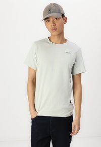 Lichtgroene T-shirt met korte mouwen en ronde hals, voorzien van een klein logo, gecombineerd met donkerdenim broek. Een gestructureerde grijze pet maakt de look compleet.