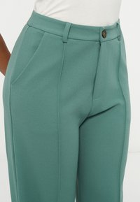 Pantalon taille haute couleur sarcelle avec passants pour ceinture, poches latérales, couture avant, et fermeture par bouton foncé, associé à un haut blanc rentré.