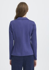 Ženska z dolgimi rjavimi lasmi, oblečena v oprijet vijoličen blazer in temno modre teksturirane hlače, prikazana s hrbtne strani na enobarvni podlagi.
