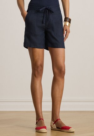 Femeie purtând pantaloni scurți bleumarin cu șnur, cu buzunare frontale, espadrile roșii și bej, și brățări aurii și de tartar pe încheietura mâinii drepte.