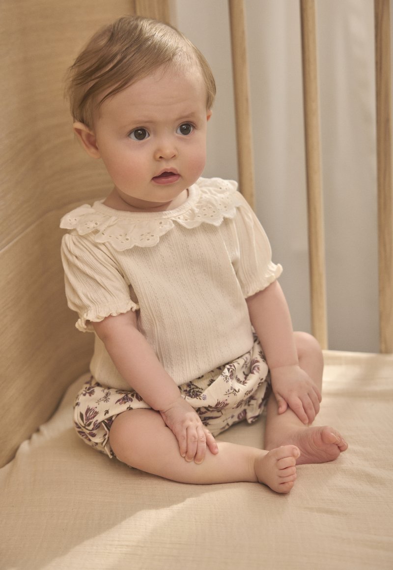 Niemowlę siedzi na beżowej poduszce wewnątrz drewnianego łóżeczka, ubrane w kremową bluzkę z koronkowym kołnierzykiem i kwieciste szorty.