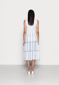 Nainen, jolla on suorat tummat hiukset, pukeutuneena valkoiseen hihattomaan toppiin, valkoiseen ja siniseen kuvioituun kerrokselliseen midi-hameeseen sekä valkoisiin paksupohjaisiin sandaaleihin, seisoo.