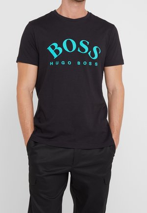 Mies pukeutuneena mustaan lyhythihaiseen T-paitaan, jossa on suuri turkoosi "BOSS HUGO BOSS" -logo, ja mustiin housuihin, seisoo käsivarsinsa taskuissa.