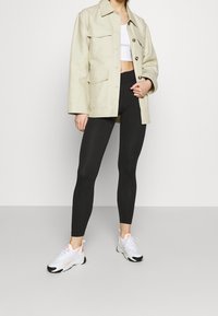 Beige arbetsjacka med stora fickor, svarta leggings och vita sneakers med orangea accenter. Släta texturer; avslappnad silhuett.