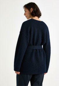 Cardigan lavorato a maglia blu navy con finitura testurizzata, maniche larghe e vita con cintura. Presenta una silhouette rilassata e una lunghezza media nella parte posteriore.