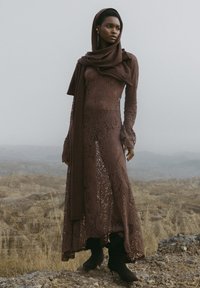 Hnedé čipkované šaty s dlhými rukávmi, rozšírenými manžetami a rozpoleným spodkom; v kombinácii s ladiacou šatkou a čiernymi členkovými čižmami. Textúrovaný kvetinový vzor.