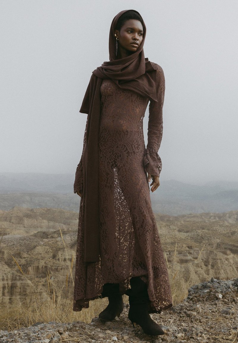 Hnedé čipkované šaty s dlhými rukávmi, rozšírenými manžetami a rozpoleným spodkom; v kombinácii s ladiacou šatkou a čiernymi členkovými čižmami. Textúrovaný kvetinový vzor.