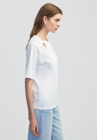 Witte katoenen t-shirt met korte mouwen, voorzien van een ronde hals en een uitsnijding op de schouder. Gecombineerd met lichtblauwe spijkerbroek.