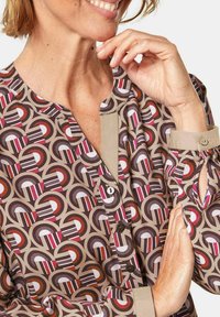 Musterbluse mit kragenförmigem Ausschnitt, die geometrische Formen in Brauntönen, Rosa und Weiß zeigt. Knopfdetails an der Vorderseite.