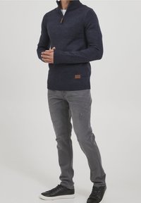 Homme portant un pull à col zippé bleu marine, un jean gris slim-fit et des baskets noires basses, se tenant les mains jointes.