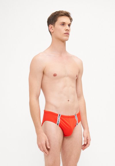 Rote Schwimmslips aus dehnbarem Material, mit kontrastierenden schwarzen Seitenstreifen und enger Passform, gezeigt an einem stehenden Modell.