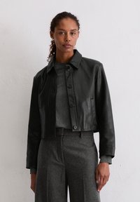 Czarna skórzana krótka kurtka z wyprofilowanym kołnierzem, zapięciem na guziki i bocznymi kieszeniami. Model ma na sobie szary sweter i dopasowane szare spodnie.