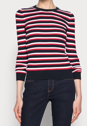 Femme portant un pull côtelé avec des rayures horizontales bleu marine, rouge et blanc, associé à un jean bleu foncé.