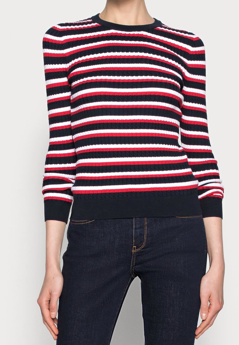 Femme portant un pull côtelé avec des rayures horizontales bleu marine, rouge et blanc, associé à un jean bleu foncé.