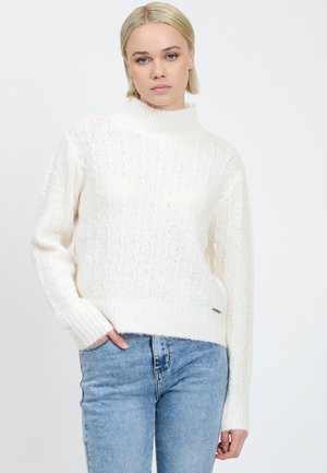 Witte trui met een hoge hals, geribde mouwen en een getextureerd kabelbreipatroon. Gecombineerd met lichtblauwe skinny jeans. Zilveren logoaccent.