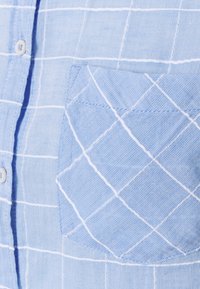 Camicia azzurro chiaro con bottone, caratterizzata da un motivo a griglia bianca, con taschino sul petto e cuciture diagonali in tessuto testurizzato.