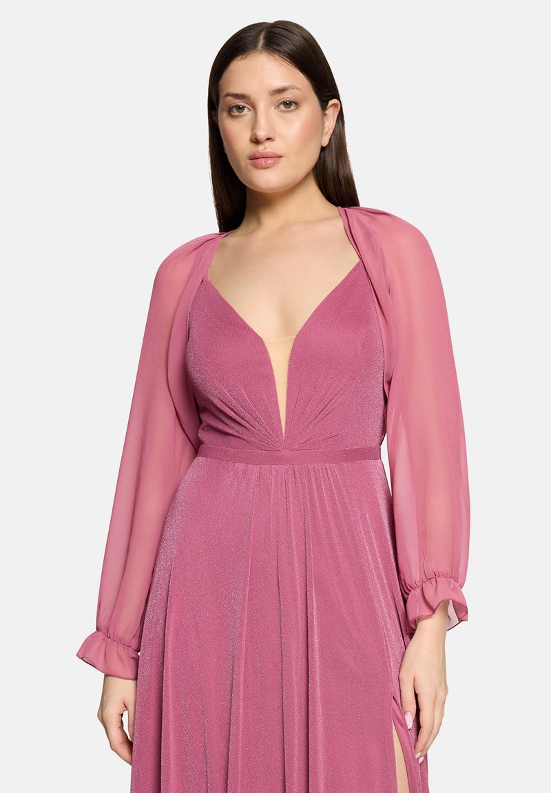 Frau mit glattem dunklem Haar, die ein tiefrosa schimmerndes Kleid mit durchsichtigen langen Ärmeln und einem hohen Schlitz an einer Seite trägt.