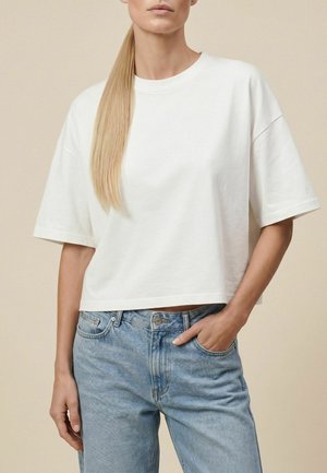 Femme aux longs cheveux blonds et raides, portant un T-shirt blanc ample et un jean bleu clair, une main dans la poche, debout devant un fond beige.