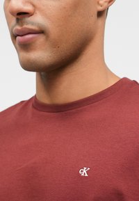 Bordový crewneck tričko vyrobené z bavlny, s malou bielou výšivkou loga "CK" na ľavej strane hrudi. Hladká textúra.