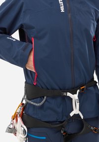 Granatowa, wodoodporna kurtka z czerwonymi akcentami zamka, z wysokim kołnierzem, bocznymi kieszeniami, noszona na uprzęży z wyposażeniem wspinaczkowym.