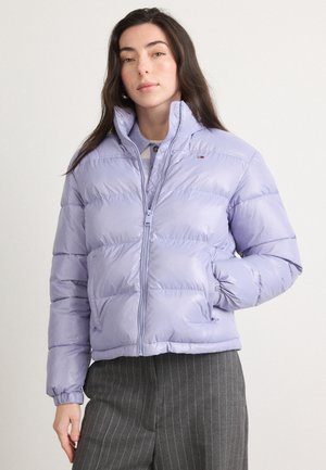 Veste remplie lavande avec un design matelassé, fermeture éclair et poignets élastiques. Portée sur une chemise bleu clair et un pantalon à rayures grises.