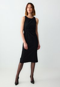 Schwarzes ärmelloses Midikleid mit einer verdrehten Frontdetails, aus weichem Stoff gefertigt, kombiniert mit durchsichtigen schwarzen Strumpfhosen und spitzen High Heels.