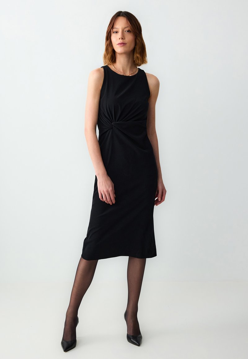 Schwarzes ärmelloses Midikleid mit einer verdrehten Frontdetails, aus weichem Stoff gefertigt, kombiniert mit durchsichtigen schwarzen Strumpfhosen und spitzen High Heels.