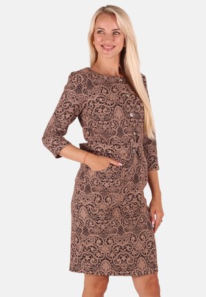 Frau mit langen blonden Haaren, die ein braunes, gemustertes knielanges Kleid mit Dreiviertelärmeln trägt, lächelt und posiert mit einer Hand in der Tasche.