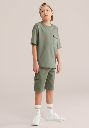 Berniukas stovintis, apsirengęs alyvuogių žalumos per dydį dideliu marškinėliu ir krovinių šortais, dėvi smėlio spalvos atvirkštinę kepurę, baltus sportbačius ir dryžuotas kojines smėlio spalvos fone.