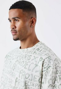 Sudadera en color crema claro con un estampado gráfico texturizado en verde de la palabra "PROJECT" en varios estilos; cuello redondo y mangas cortas.