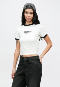 Νεαρή γυναίκα που φοράει λευκό κοντομάνικο τοπ με μαύρο περίγραμμα, μαύρα τζην, γκρι καπέλο φοράει ανάποδα και έχει τατουάζ στο αριστερό της χέρι, στέκεται με το ένα χέρι στην τσέπη.