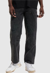 Black denim cargo pants featuring a relaxed fit, distressed accents, and multiple pockets. Worn with white sneakers for contrast.