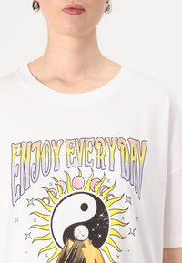 White cotton t-shirt featuring a colorful graphic design with "ENJOY EVERY DAY," a yin-yang symbol, sun rays, and stars in a vintage style.