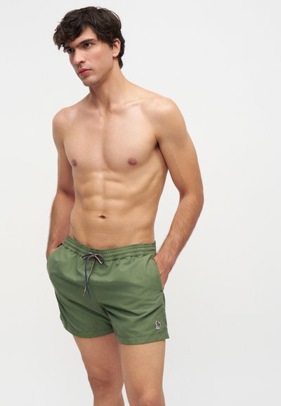 Jeune homme aux cheveux courts et foncés portant un short de bain vert, les mains dans les poches, sur un fond clair uni.