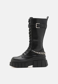 Schwarzer knielanger Kampf-Stiefel mit einer klobigen Plattformsohle, Schnürvorderteil, silberner Kettenakzent und verstellbarem Riemen. Glatte Lederoberfläche.