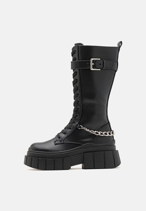 Bottes de combat noires montant jusqu'au genou avec une semelle plateforme épaisse, un laçage à l'avant, un accent en chaîne argentée et une sangle à boucle ajustable. Fini en cuir lisse.