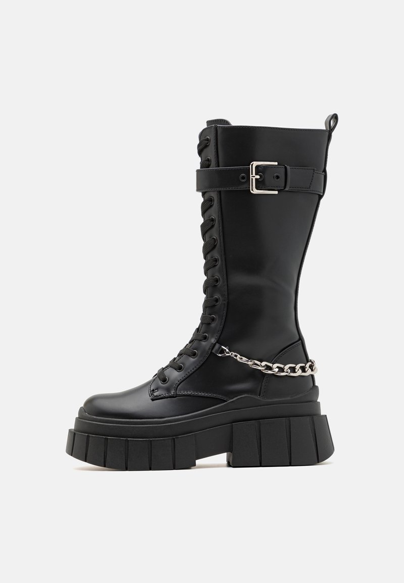 Schwarzer knielanger Kampf-Stiefel mit einer klobigen Plattformsohle, Schnürvorderteil, silberner Kettenakzent und verstellbarem Riemen. Glatte Lederoberfläche.