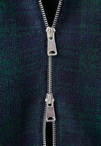 Sweter z suwakiem w kolorze granatowym i zielonym, z charakterystycznym wzorem w kratę. Srebrny metalowy suwak z podwójnymi uchwytami biegnie wzdłuż przodu.