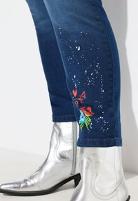 Vaqueros de denim con un lavado azul, que presentan salpicaduras blancas y bordados florales coloridos en la parte baja de la pierna, combinados con botas de vaquero plateadas.