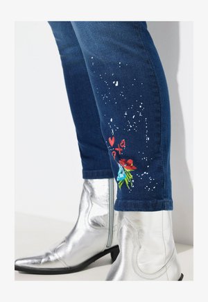 Spijkerbroek met een blauwe wassing met witte spatten en kleurrijke bloemenborduursels op de onderbenen, gecombineerd met zilveren cowboylaarzen.