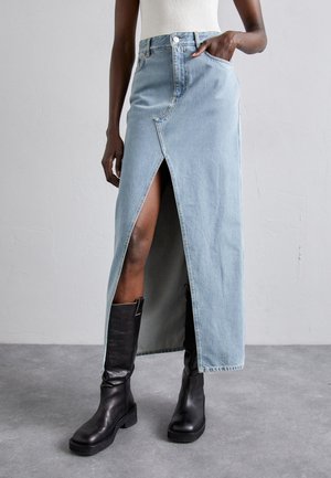 Kobieta w jasnoniebieskiej długiej spódnicy dżinsowej z przednim rozcięciem i czarnych kozakach sięgających kolan, stoi na szarej podłodze na tle jednolitej ściany.