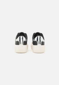 Zapatillas en blanco y negro con una suela de goma texturizada, detalles a rayas en el talón y "TOMMY" grabado en la parte trasera.